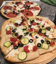 In the foreground, oven pizza with sliced zuncchini, olives, onions and mozzarella. In the background, Breville pizza with fresh sliced tomatoes, olives, mozzarella, and basil.